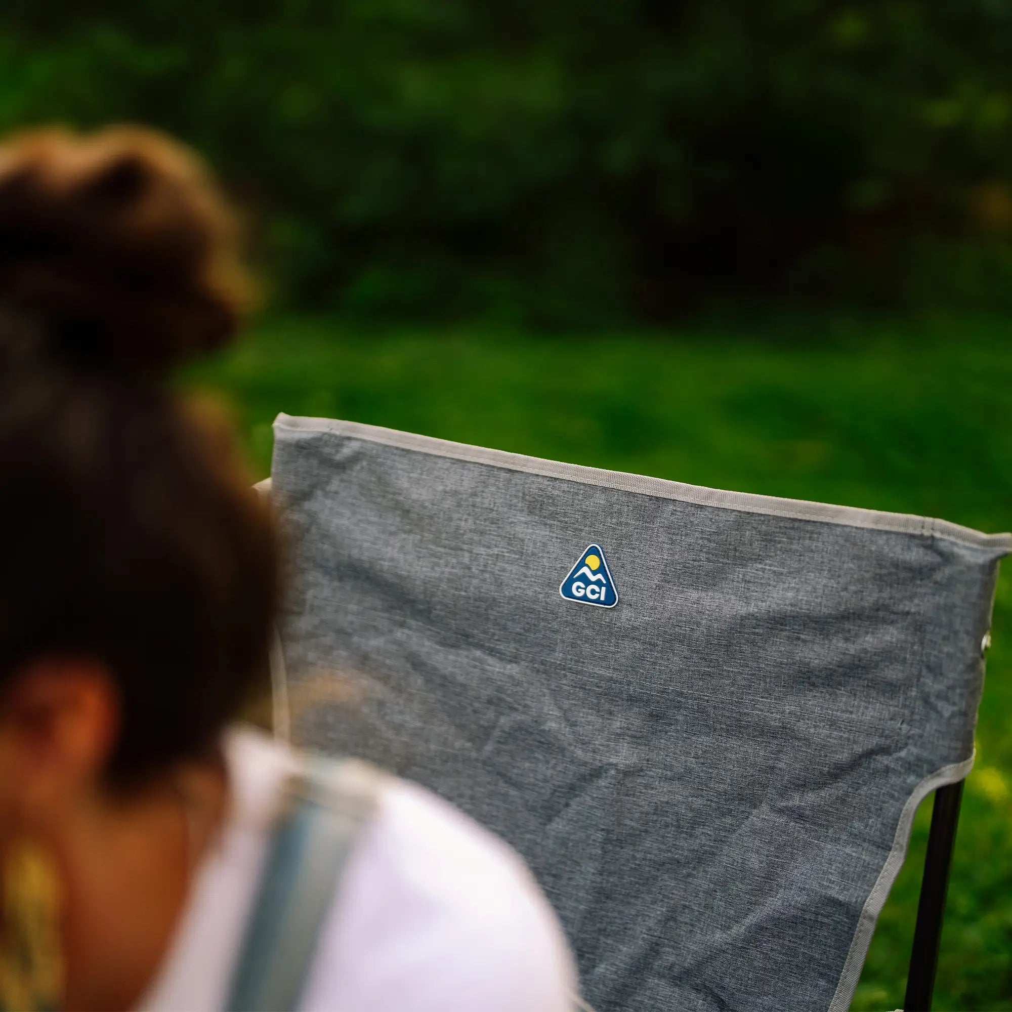 A gray Kickback Rocker with a small blue and yellow GCI logo on the backrest, photographed outdoors with a blurred green background and part of a person in the foreground.