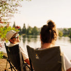 A man and a woman sit in Kickback Rockers by a lake at sunset, viewed from behind them. 