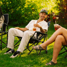A man sitting in a Kickback Rocker on a grassy lawn, holding an iced drink with a straw. He is wearing a white polo shirt, light beige pants, and a green and white baseball cap with sunglasses.