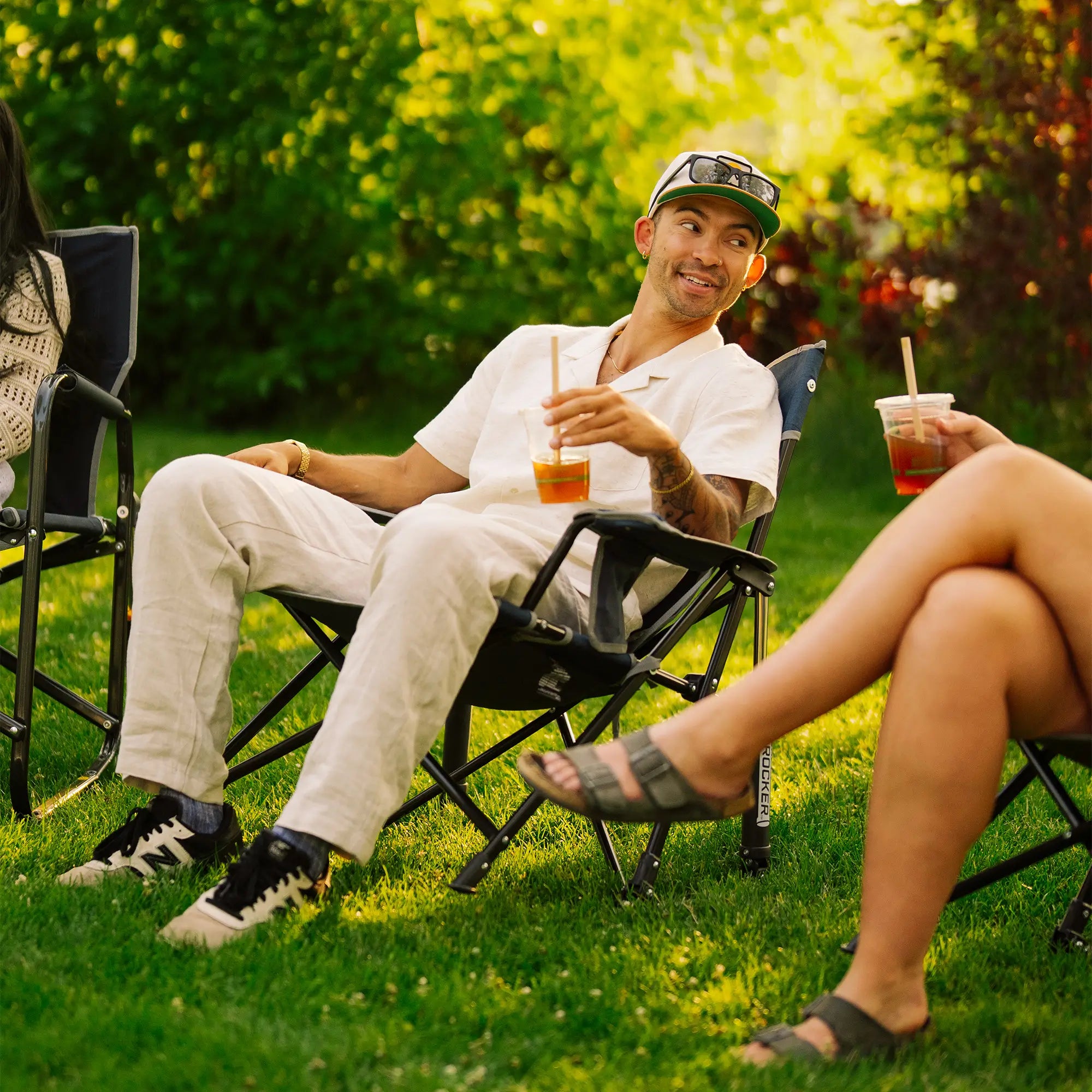 A man sitting in a Kickback Rocker on a grassy lawn, holding an iced drink with a straw. He is wearing a white polo shirt, light beige pants, and a green and white baseball cap with sunglasses.