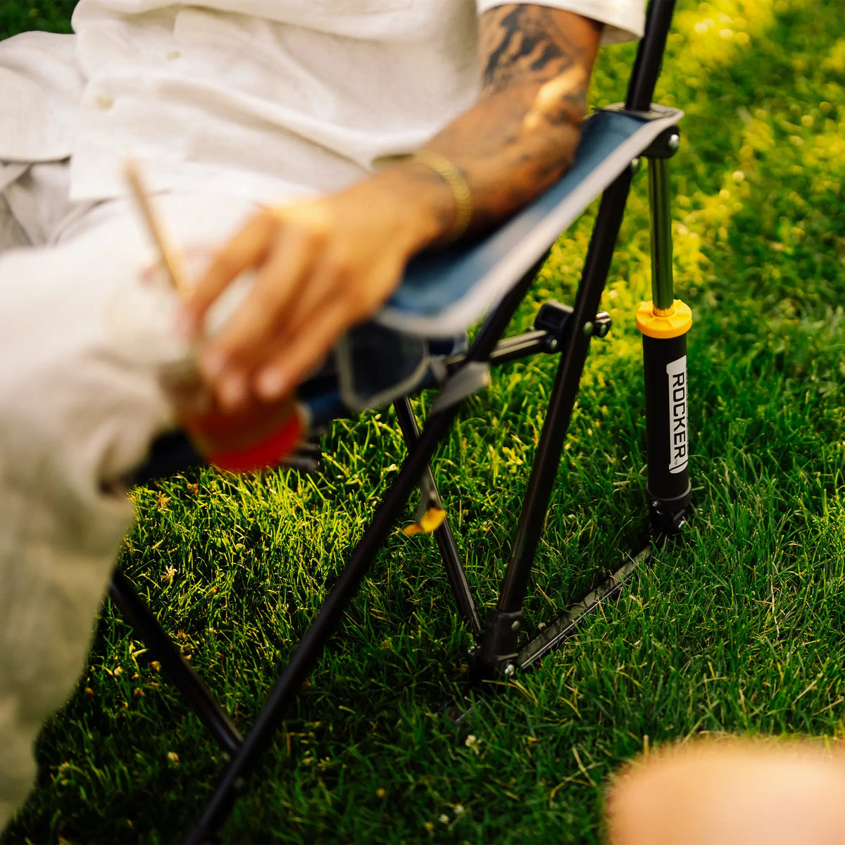 A close-up outdoor shot of a person sitting on a Kickback Rocker on grass, holding a drink. The chair has a blue fabric seat and black metal frame.