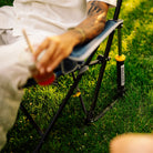 A close-up outdoor shot of a person sitting on a Kickback Rocker on grass, holding a drink. The chair has a blue fabric seat and black metal frame.