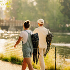 A man and a woman walk away along a sunlit path beside a lake, each carrying a folded kickback rocker on their backs. The scene is outdoors with trees, water, and warm backlighting.
