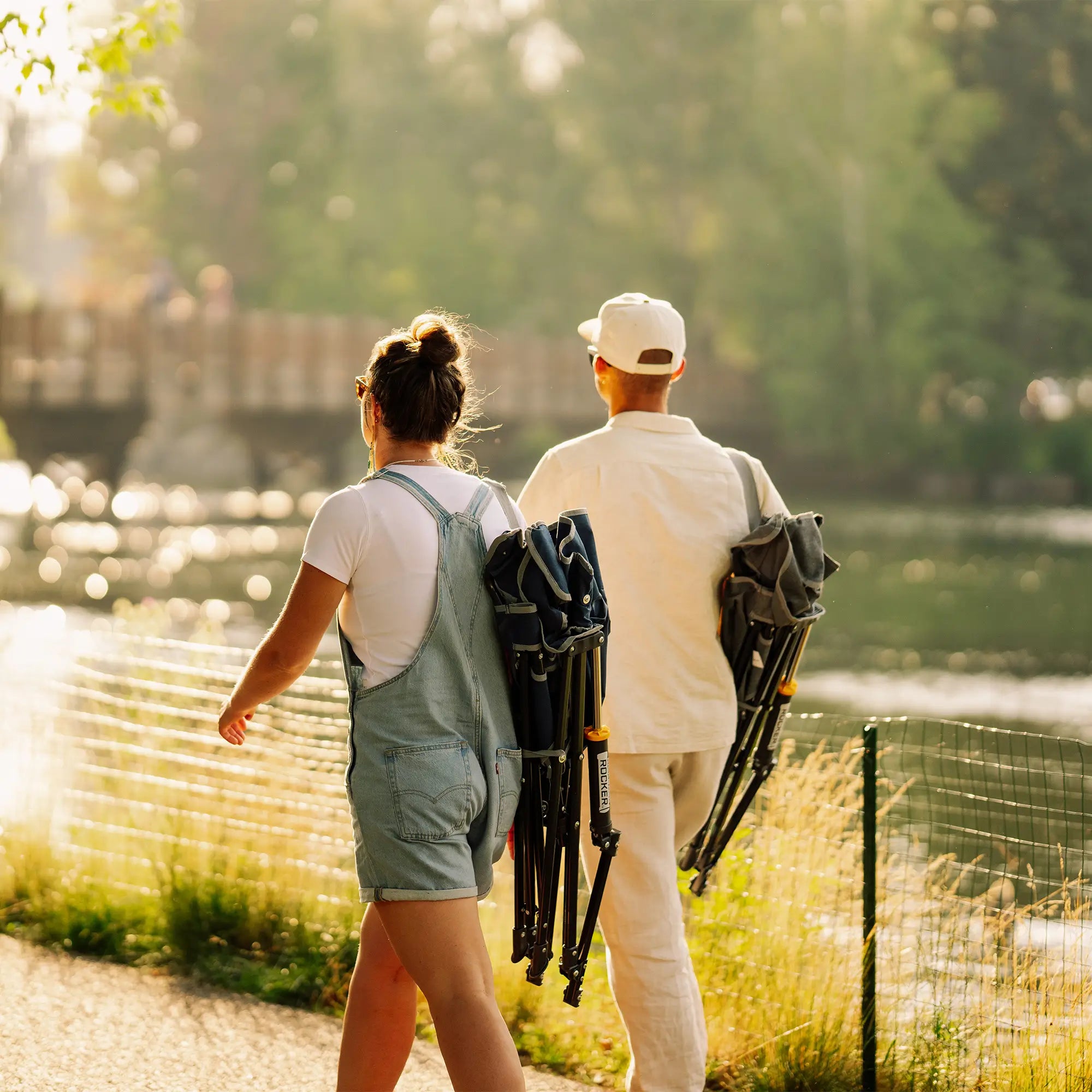 A man and a woman walk away along a sunlit path beside a lake, each carrying a folded kickback rocker on their backs. The scene is outdoors with trees, water, and warm backlighting.