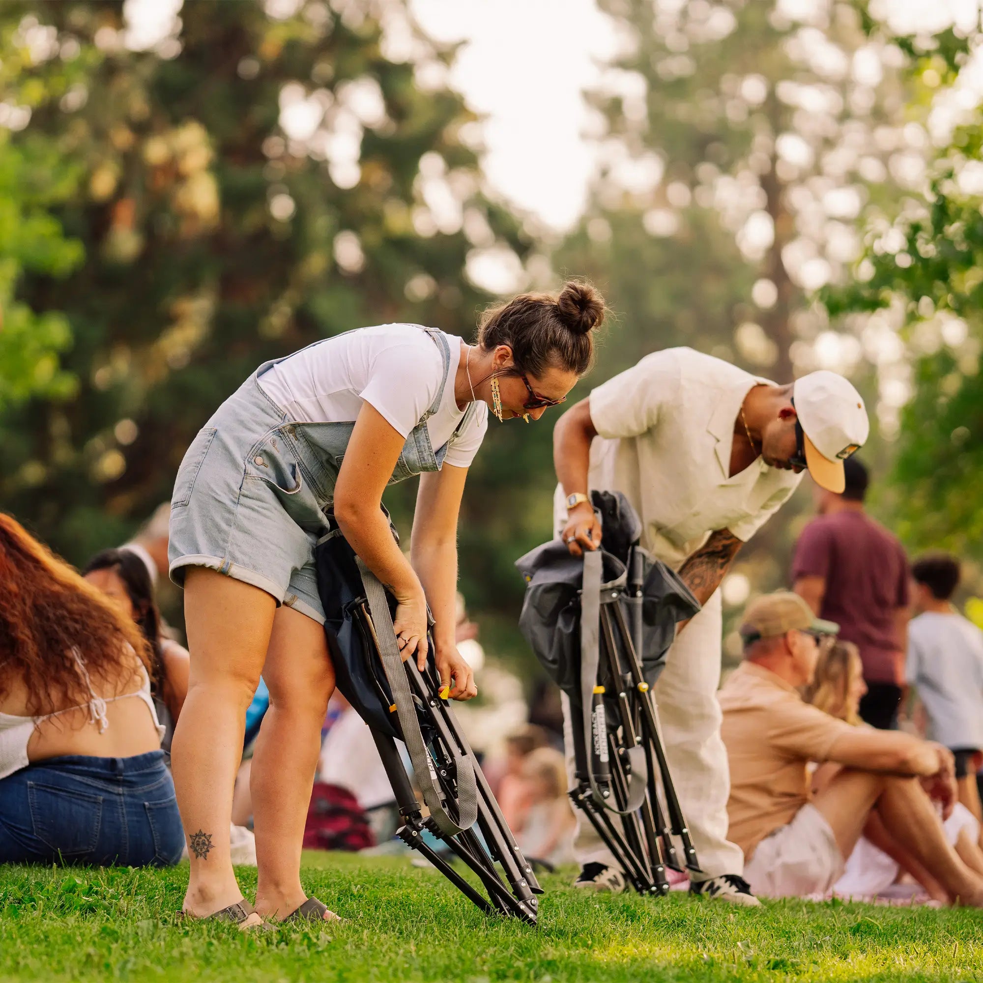 A man and a woman in a park setting, folding kickback rockers on the grass while other people relax in the background.