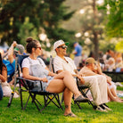 A couple sitting in indigo and gray kickback rockers on a grassy lawn at an outdoor concert or festival, with other people and trees blurred in the background.