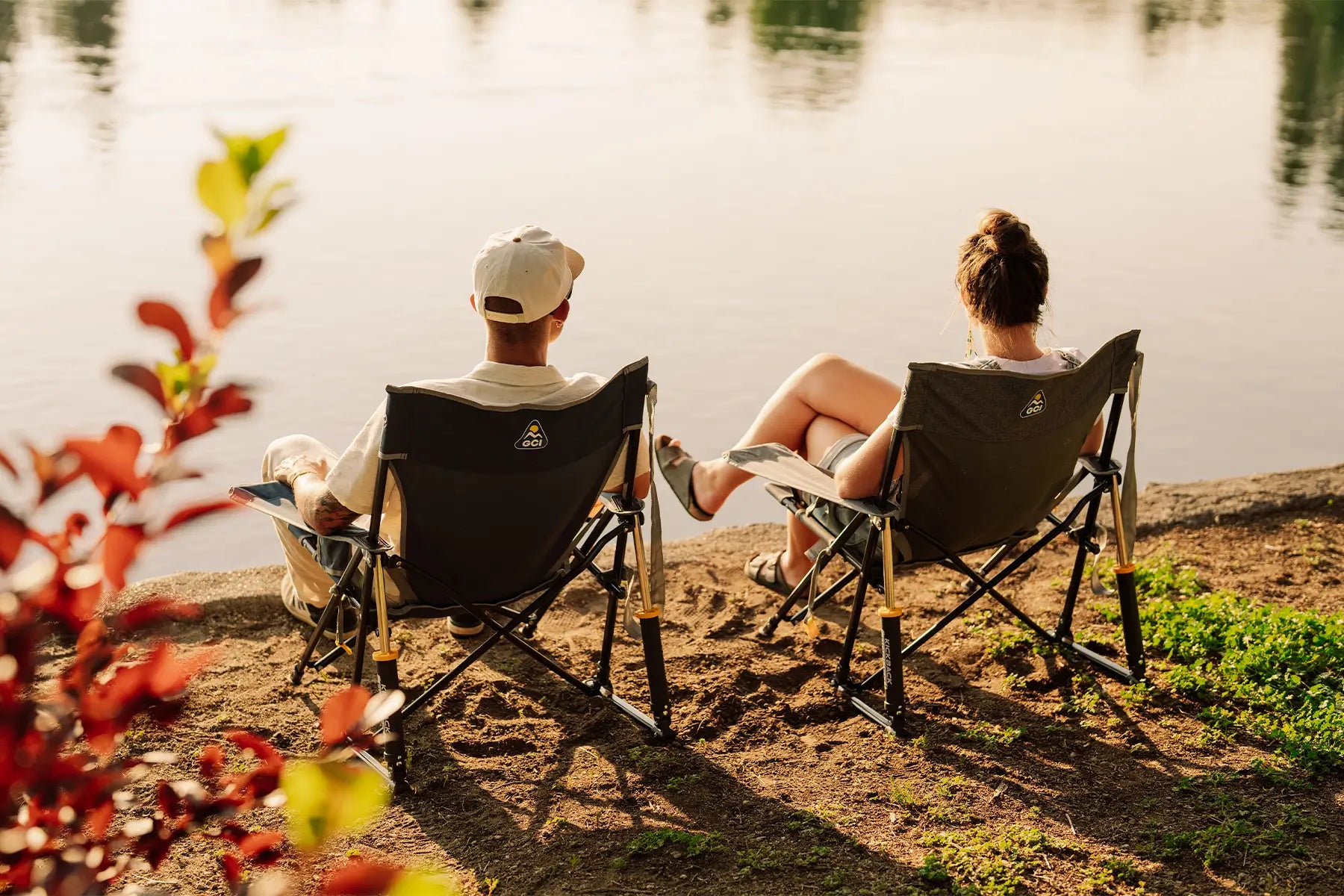 Two people sitting in Kickback Rockers on a sandy lakeshore, viewed from behind as they face the water. Red foliage is blurred in the foreground, and the warm sunlight casts long shadows on the ground.