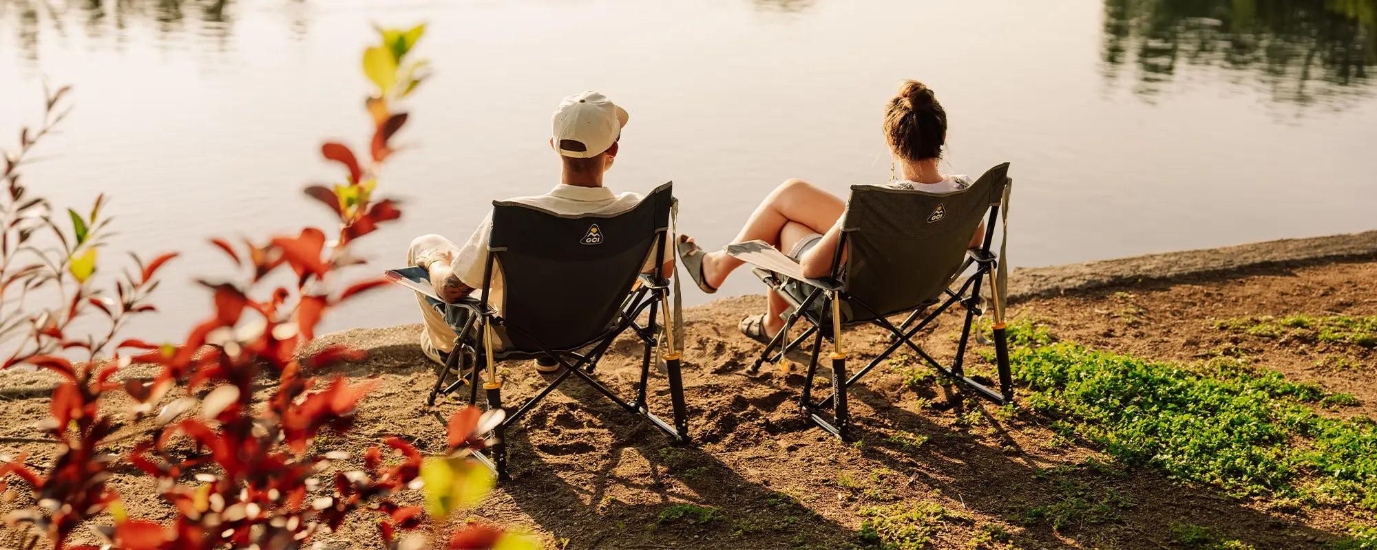 Two people sitting in Kickback Rockers on a sandy lakeshore, viewed from behind as they face the water. Red foliage is blurred in the foreground, and the warm sunlight casts long shadows on the ground.