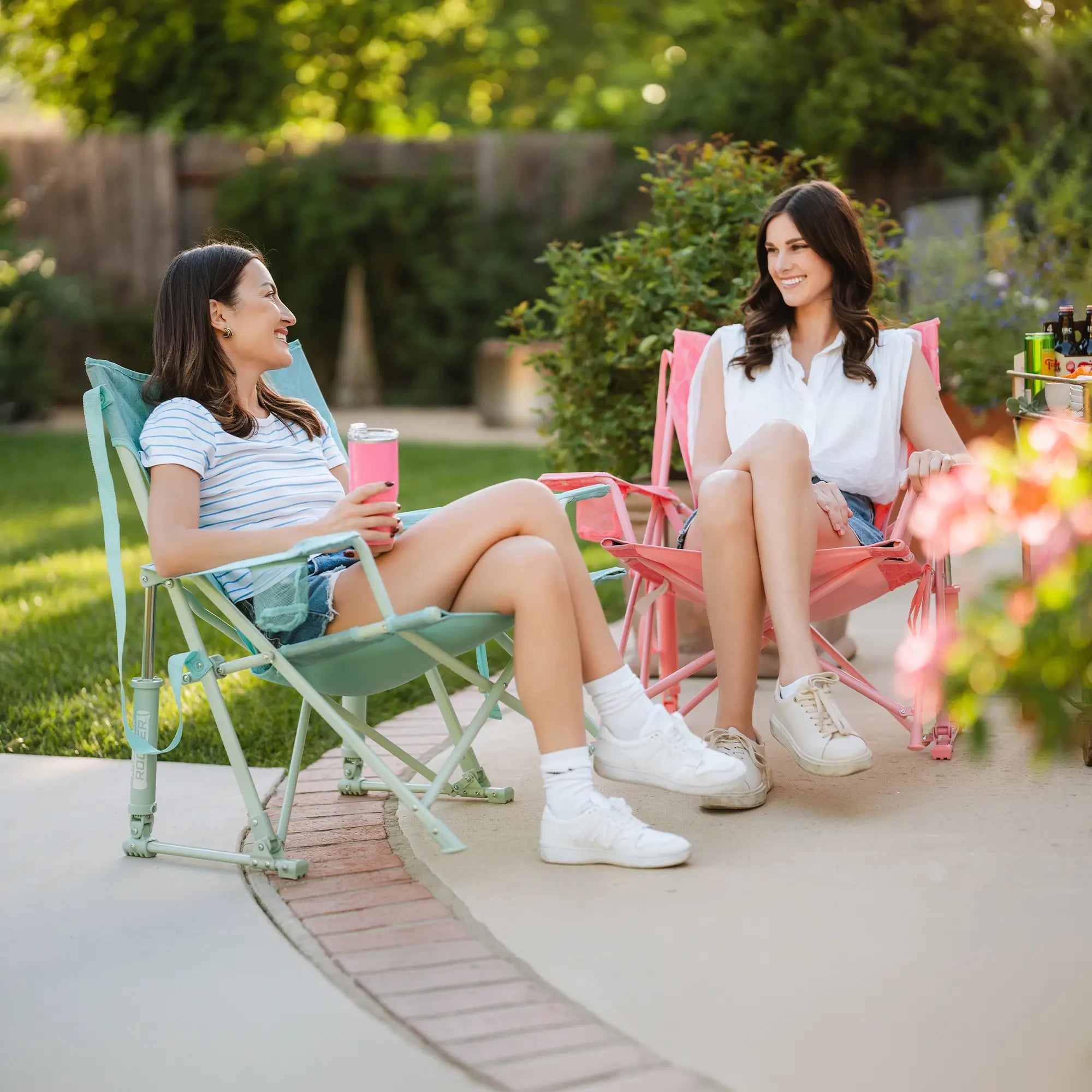Two women sitting in folding lawn chairs on a patio in a backyard setting, talking and smiling. One chair is mint green and the other is coral pink, with grass and garden foliage in the background.