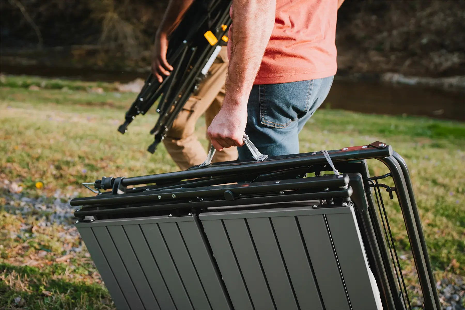 A man walking and carrying a slim-fold table next to a creek bed. 