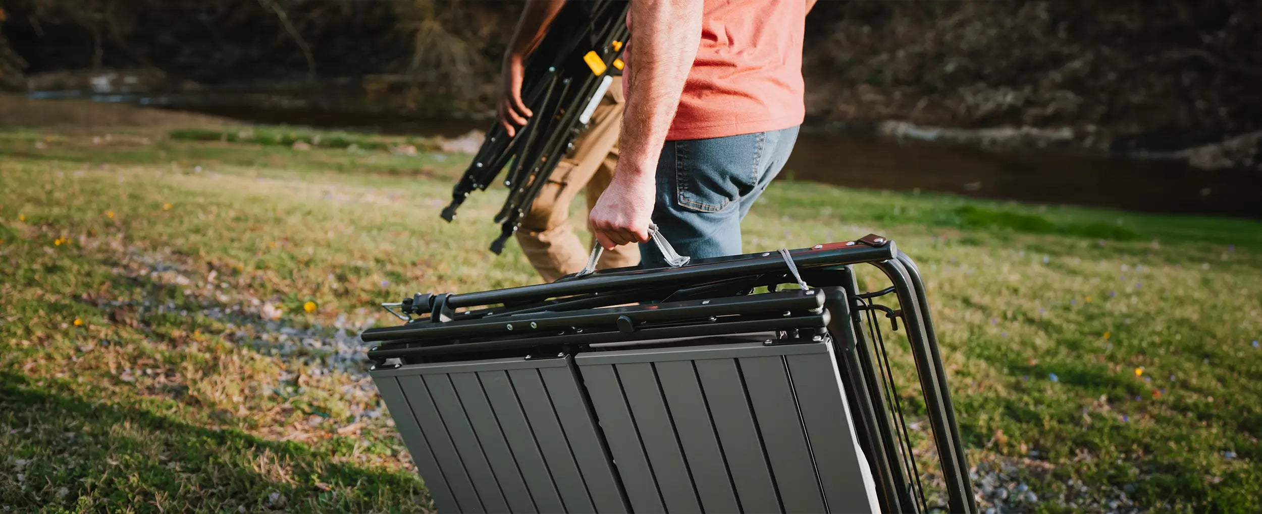 A man walking and carrying a slim-fold table next to a creek bed. 