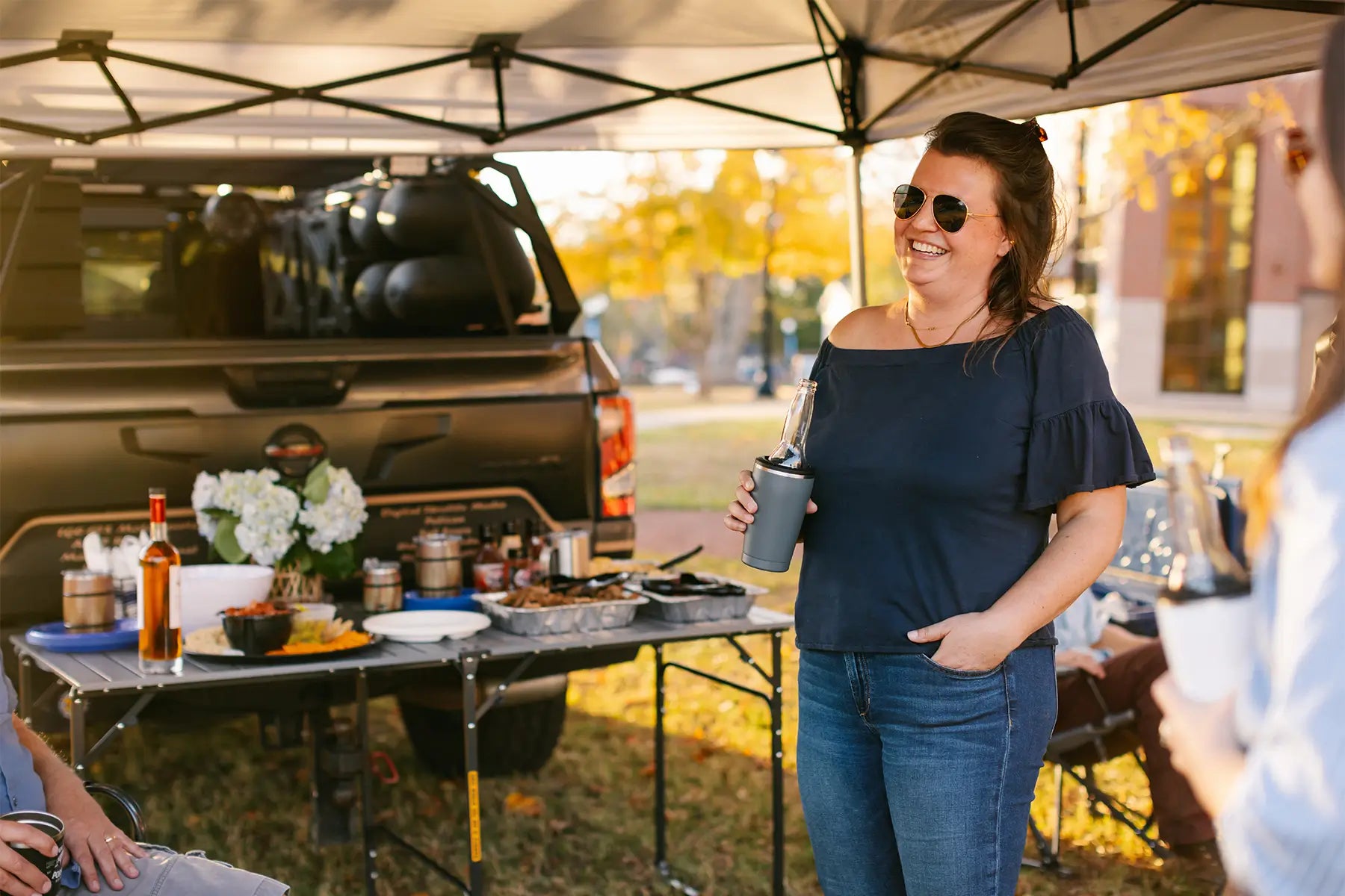A woman sitting underneath a levrup canopy at a tailgate while postioned next to a slim fold table. 