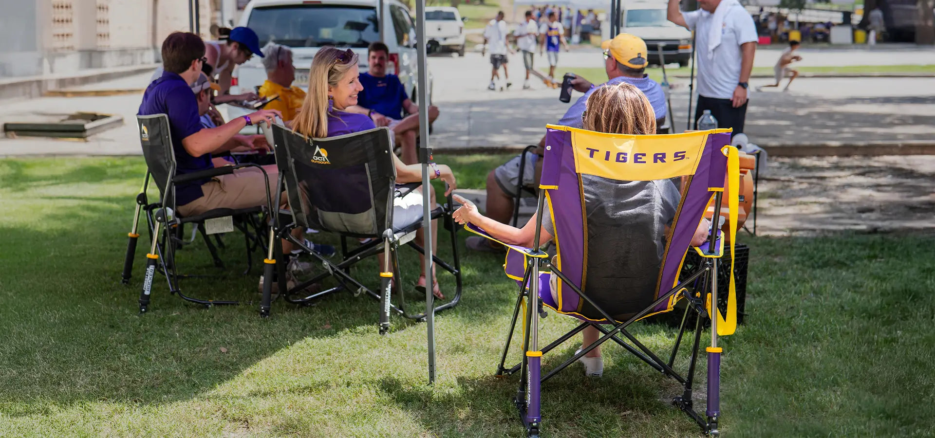 A woman sitting in an LSU comfort pro rocker that says tigers on the back of it, next to a group of tailgaters sitting under a canopy. 