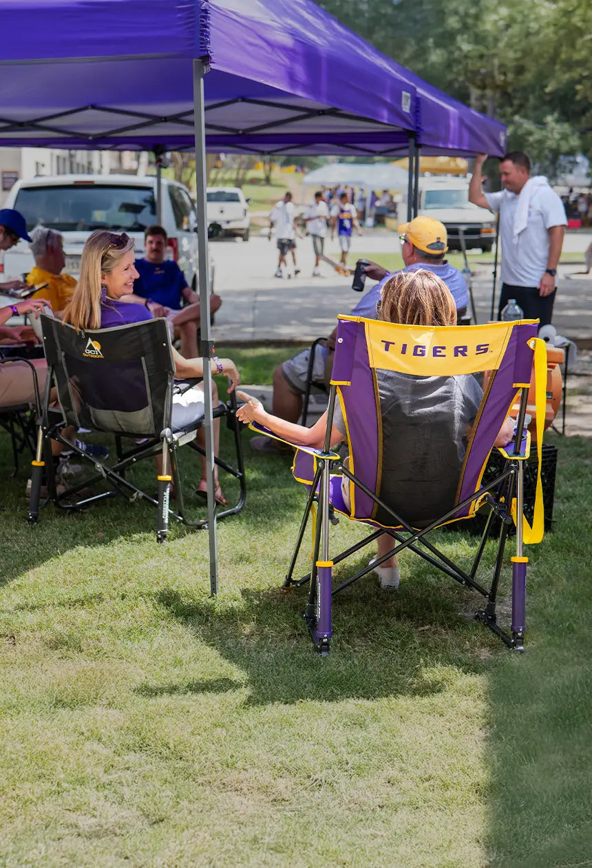 A woman sitting in an LSU comfort pro rocker that says tigers on the back of it, next to a group of tailgaters sitting under a canopy. 