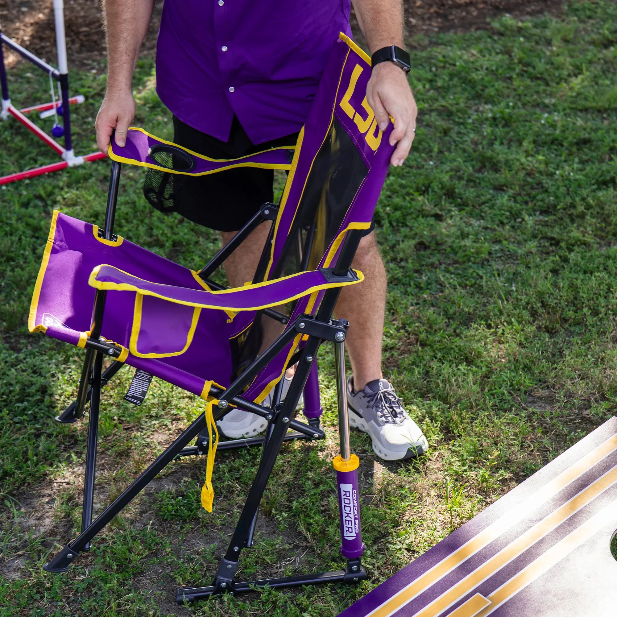A person collapsing the LSU comfort pro rocker chair next to an LSU cornhole board. 