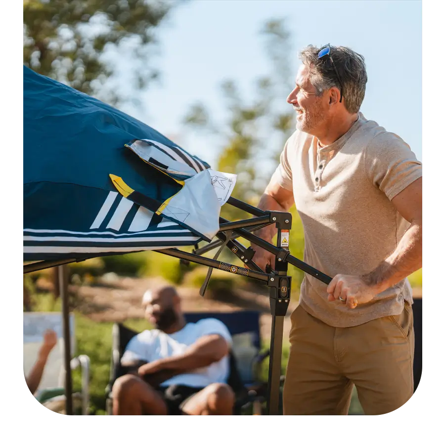 Man setting up a Levrup Shade Canopy at a sunny outdoor event with others relaxing in the background.