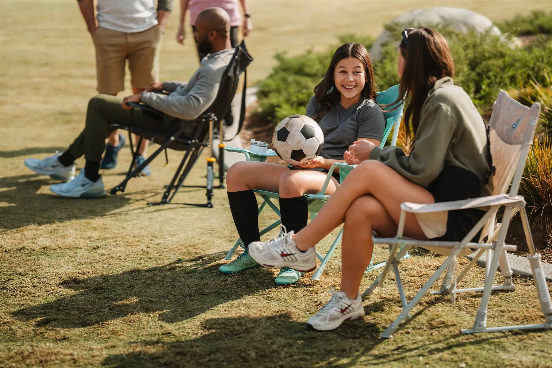A young girl and a woman talking to one another while sitting in Kickback Rocker chairs on a soccer field. 