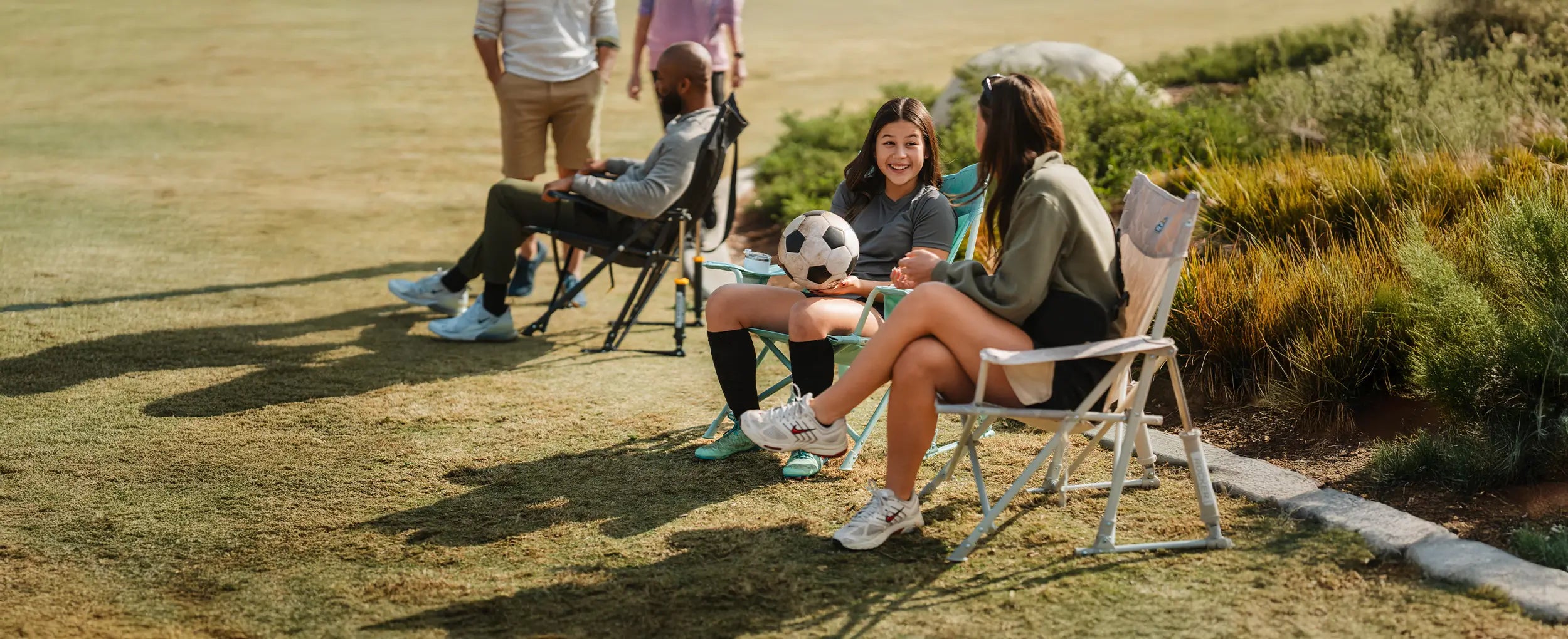 A young girl and a woman talking to one another while sitting in Kickback Rocker chairs on a soccer field. 