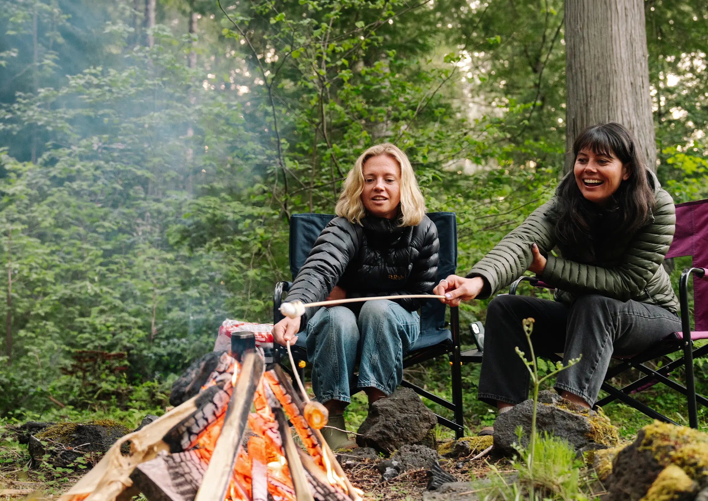 Two people sitting around a campfire in a forest, roasting marshmallows on sticks in GCI Outdoor chairs. 