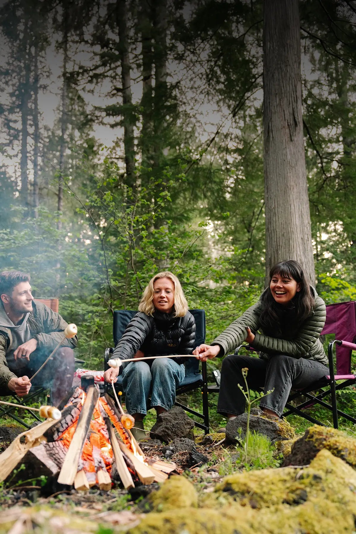 Three people sitting around a campfire in a forest, roasting marshmallows on sticks in GCI Outdoor chairs. 