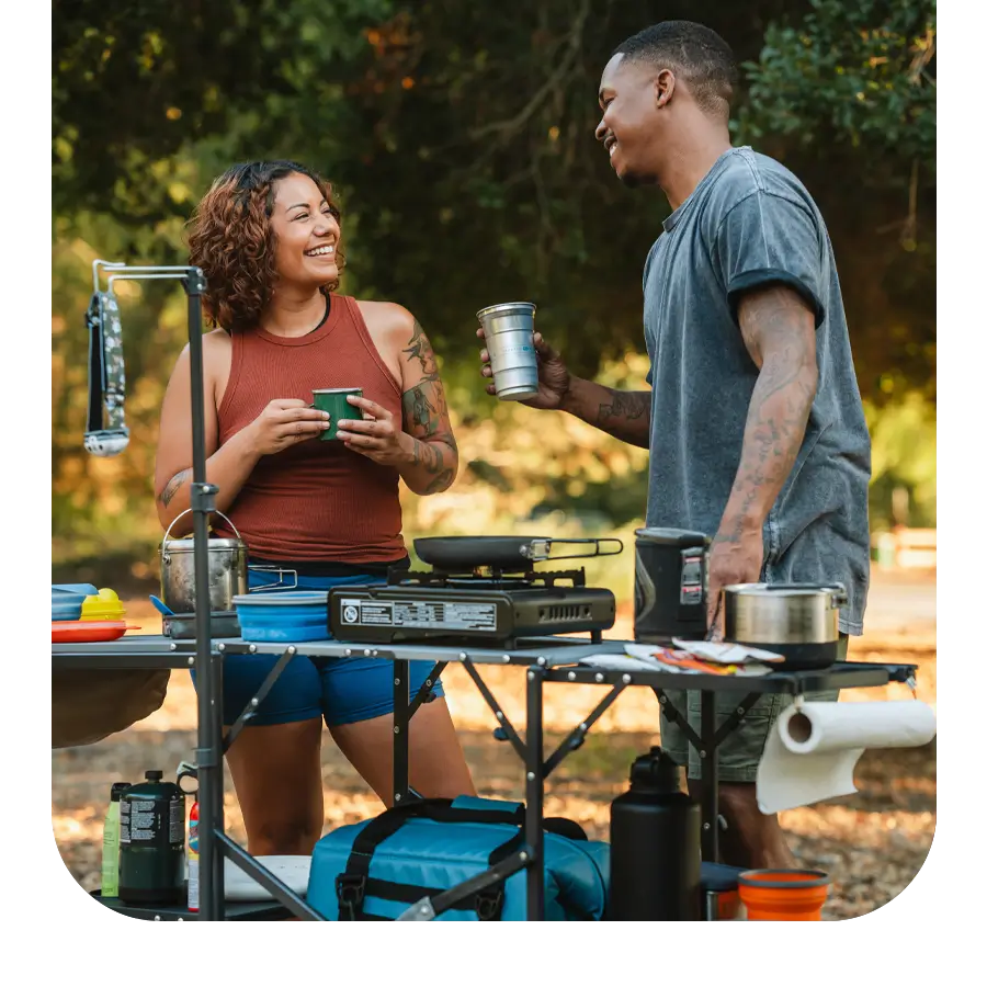 A couple enjoying drinks and conversation at a Master Cook Station set up in a scenic campsite.