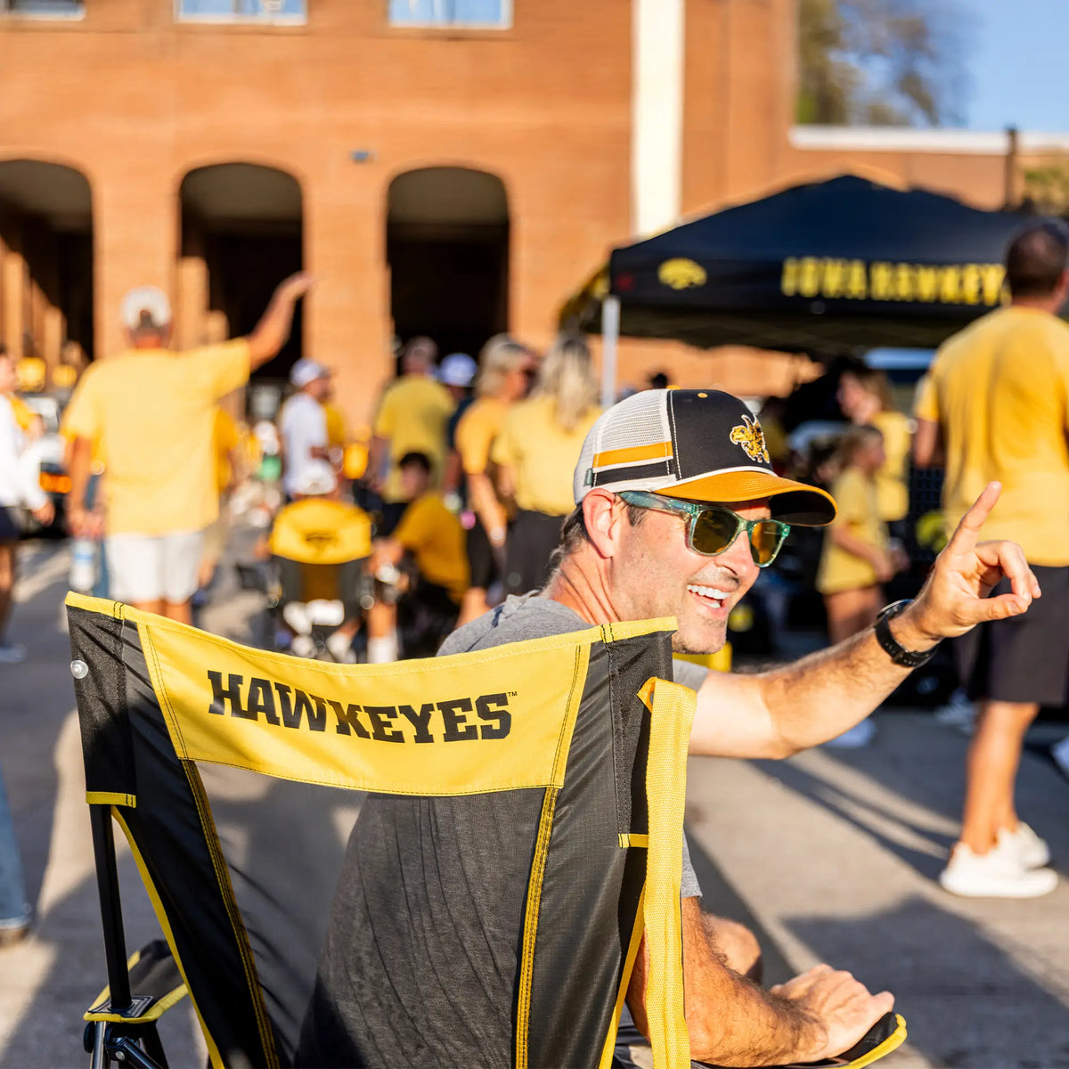 A man raising his hand to wave at somebody while sitting in an iowa state comfort pro rocker with "hawkeyes" text on the back of his chair. 
