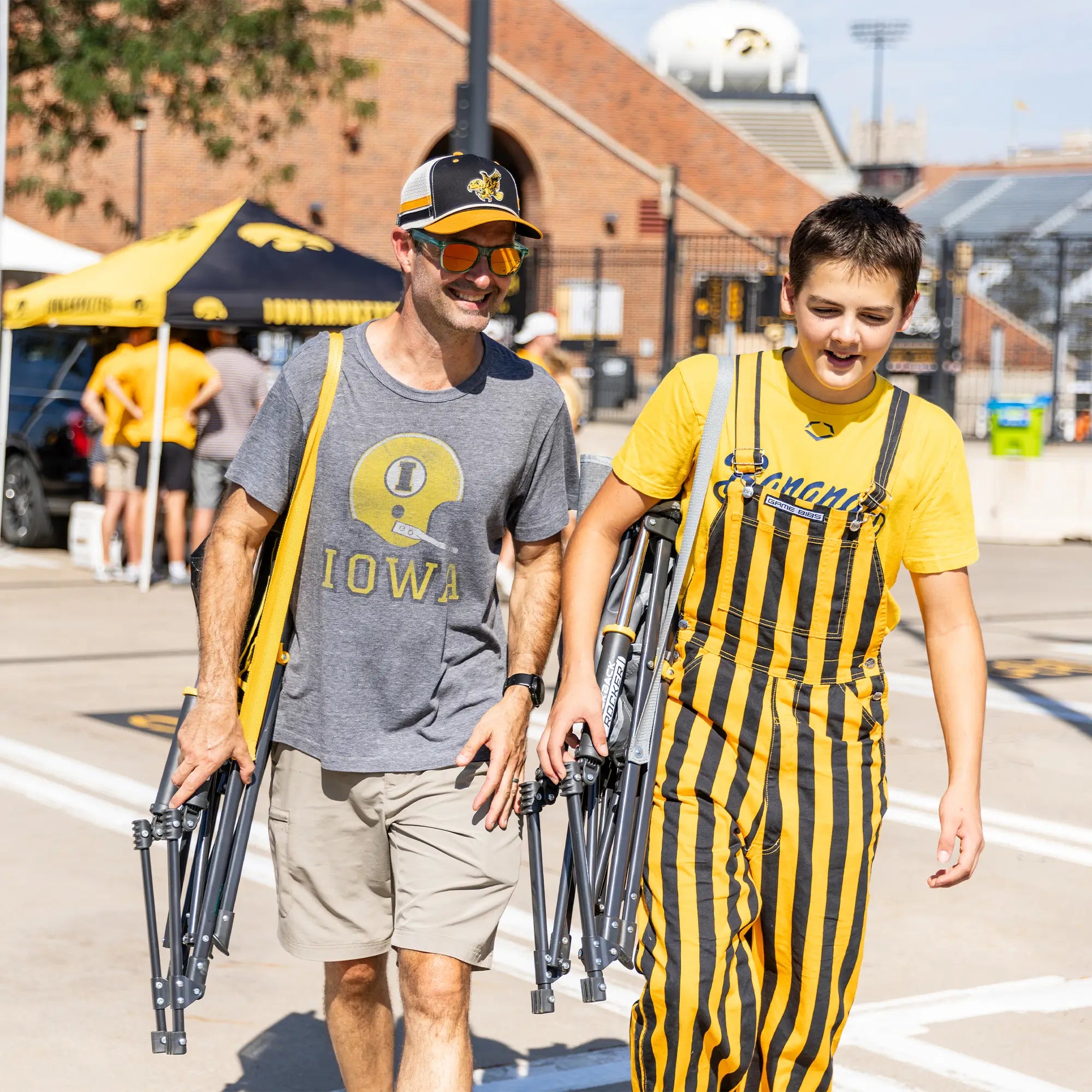 A parent and a student carrying their iowa state comfort pro rockers on their shoulder while walking through a tailgate party. 
