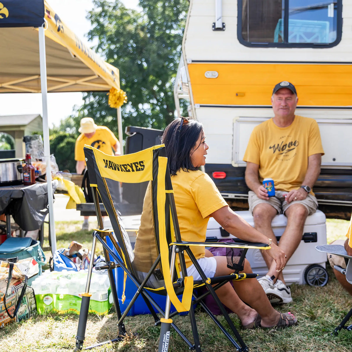 A woman sitting in an iowa state comfort pro rocker talking to a man sitting on a cooler next to a camper. 