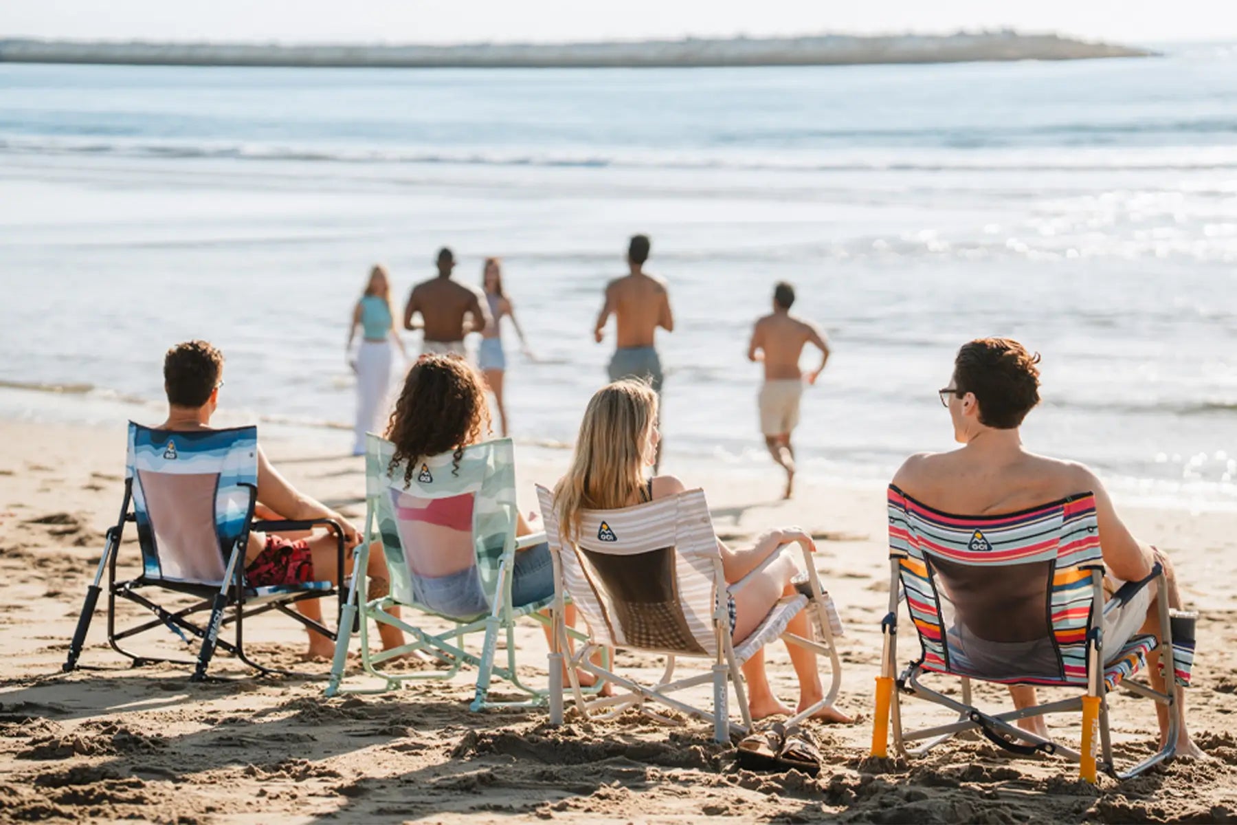 Group of friends sitting on colorful beach chairs facing the ocean, watching others play in the water.