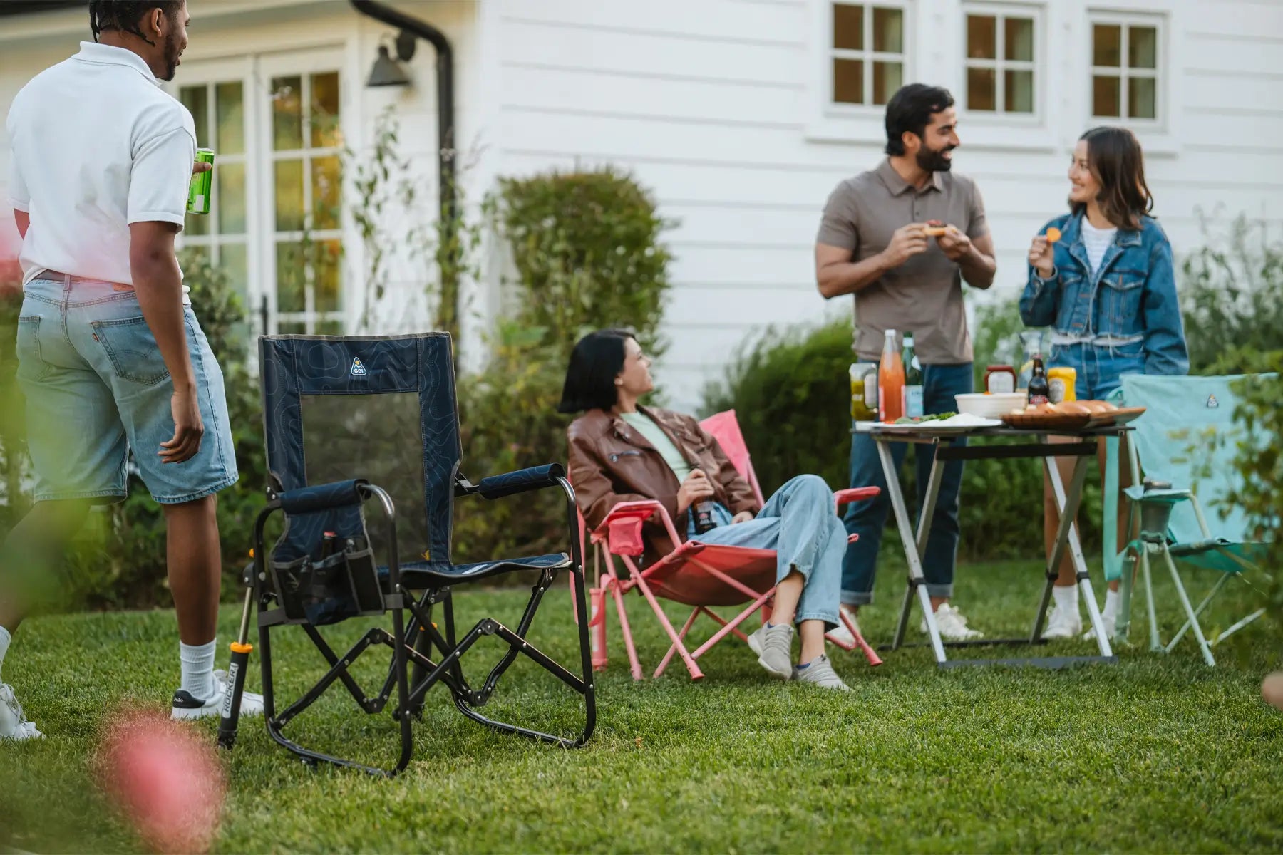 A group of friends enjoys an outdoor gathering with foldable camping chairs, a picnic table with food, and a relaxed backyard setting.