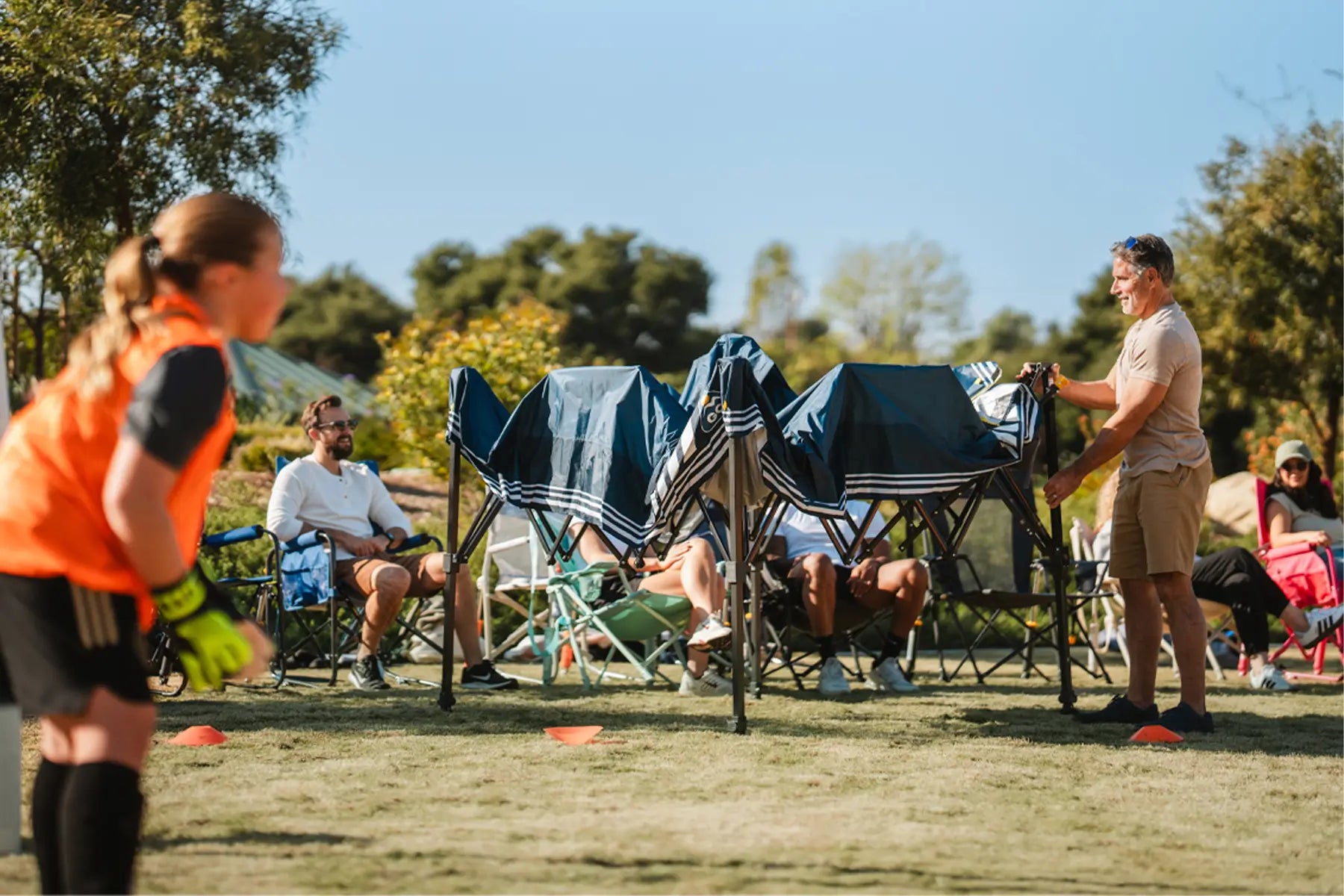 Man setting up the Levrup Shade Canopy in front of a group sitting in chairs watching a soccer game. 