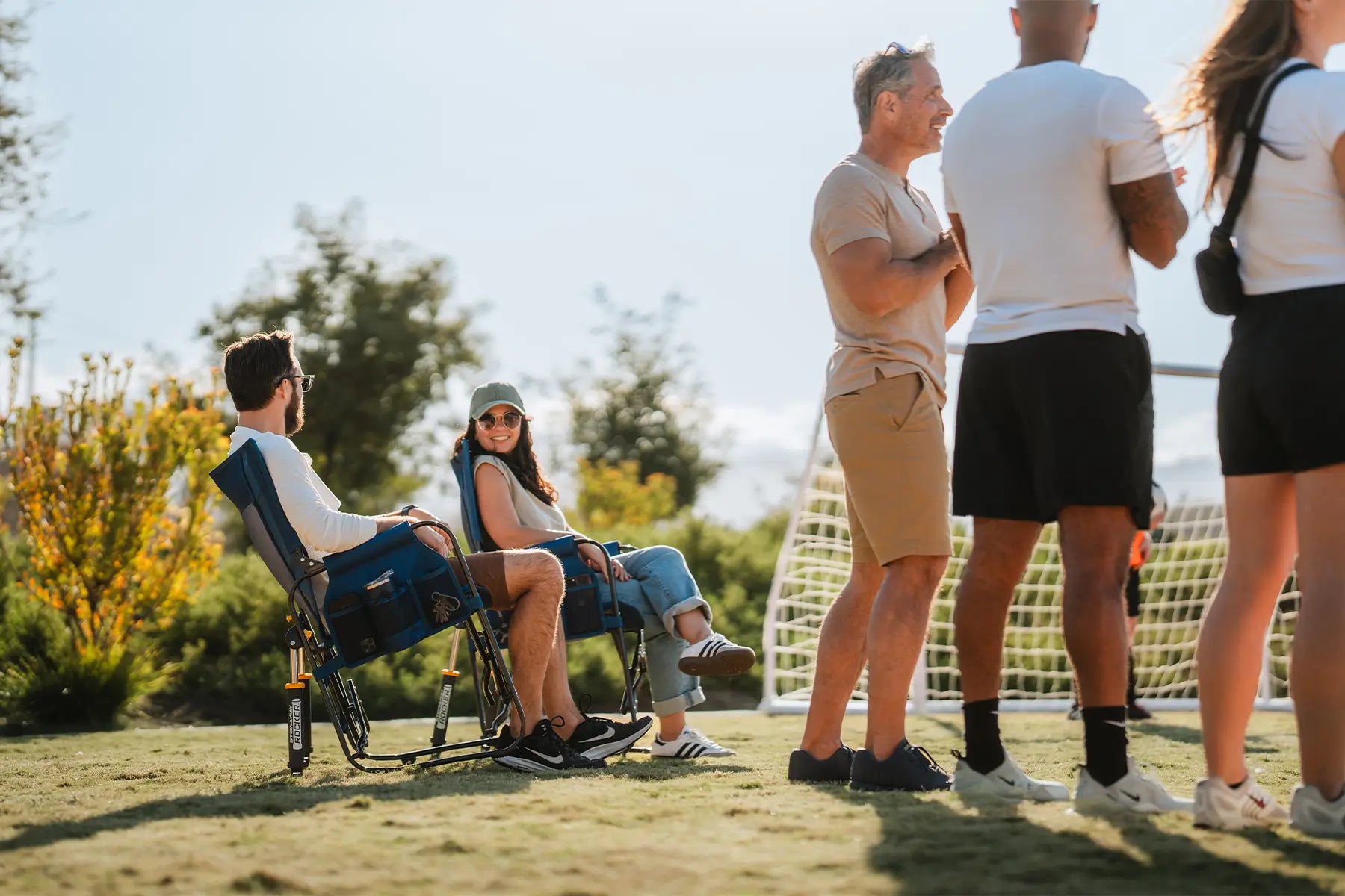 A couple laughing and sitting in two denim Stowaway Rockers next to a soccer goal net. 