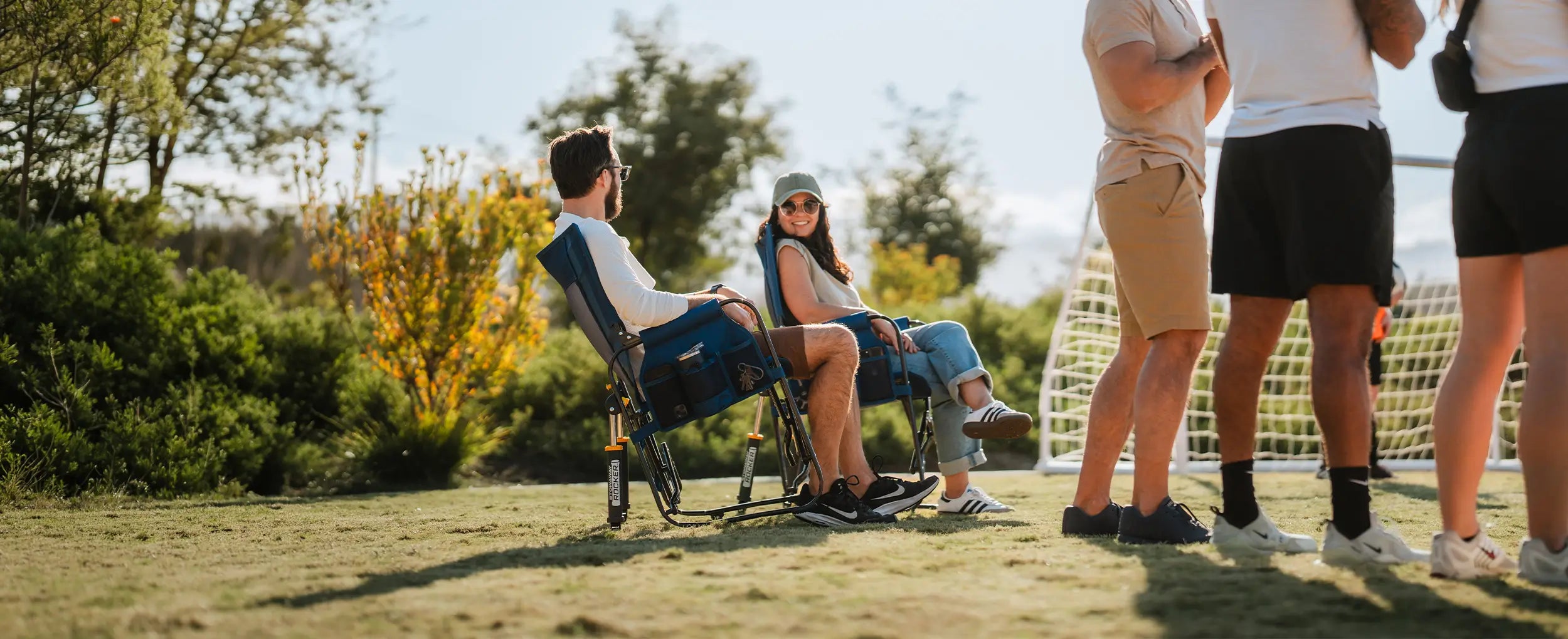 A couple laughing and sitting in two denim Stowaway Rockers next to a soccer goal net. 