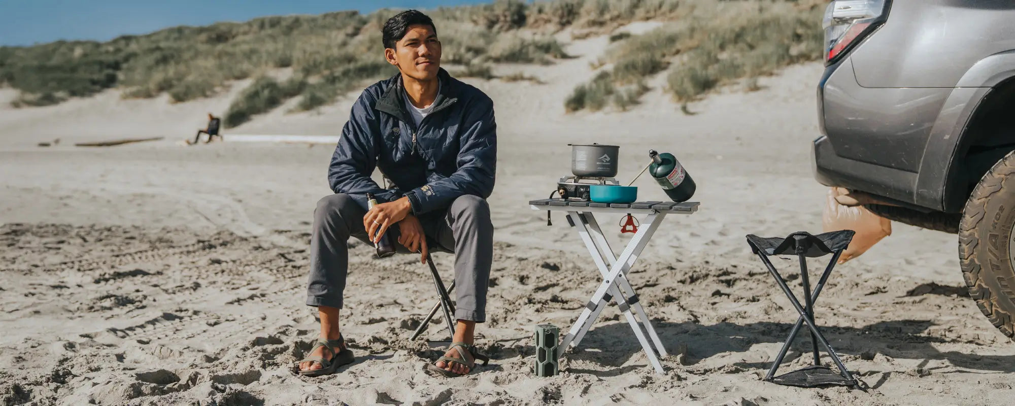 Man sitting on a Packseat on a sandy beach next to a small table with cooking gear.