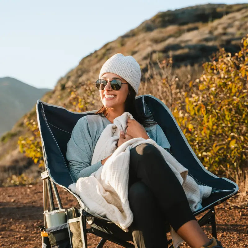 Woman bundled in a blanket, smiling and seated comfortably in a Pod Rocker chair on a scenic hillside trail.