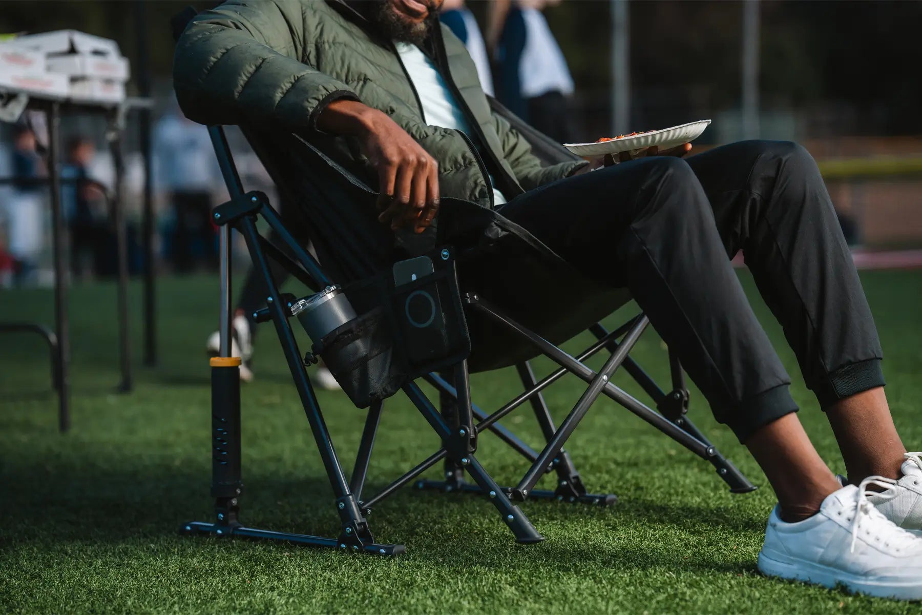 Person sitting in a Pod Rocker at a sporting event, holding a plate of food with a cup holder attached.