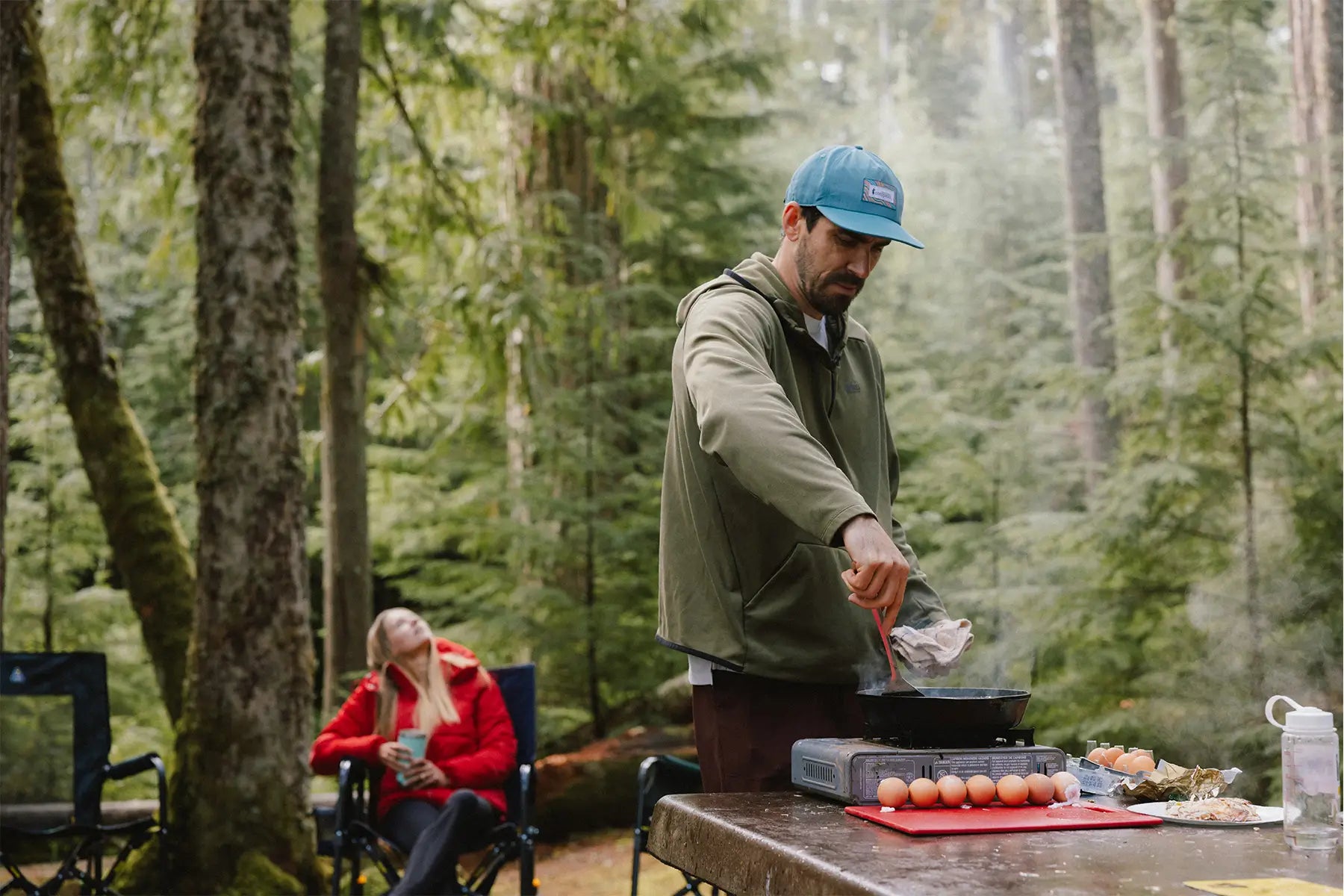 Man cooking outdoors in a forest setting with a woman sitting in the background.
