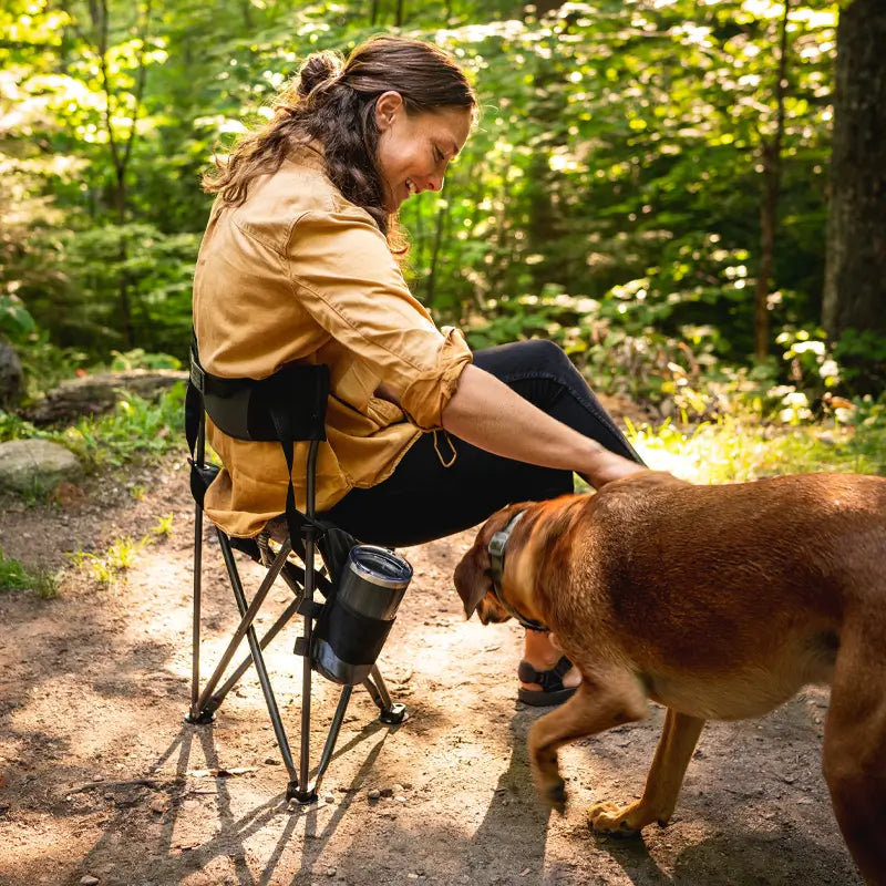 Person sitting on a black Quik-E-Seat with a drink in the cup holder, smiling and petting a dog on a dirt trail in the forest.