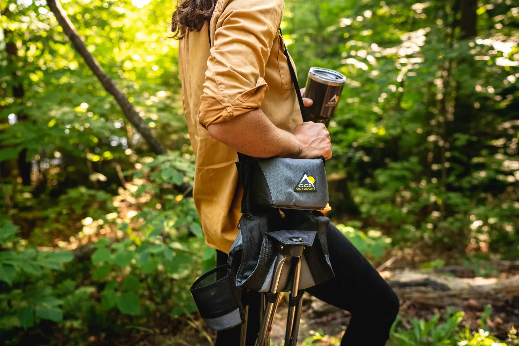 Person carrying a Quik-E-Seat through a forest, holding a branded travel mug.