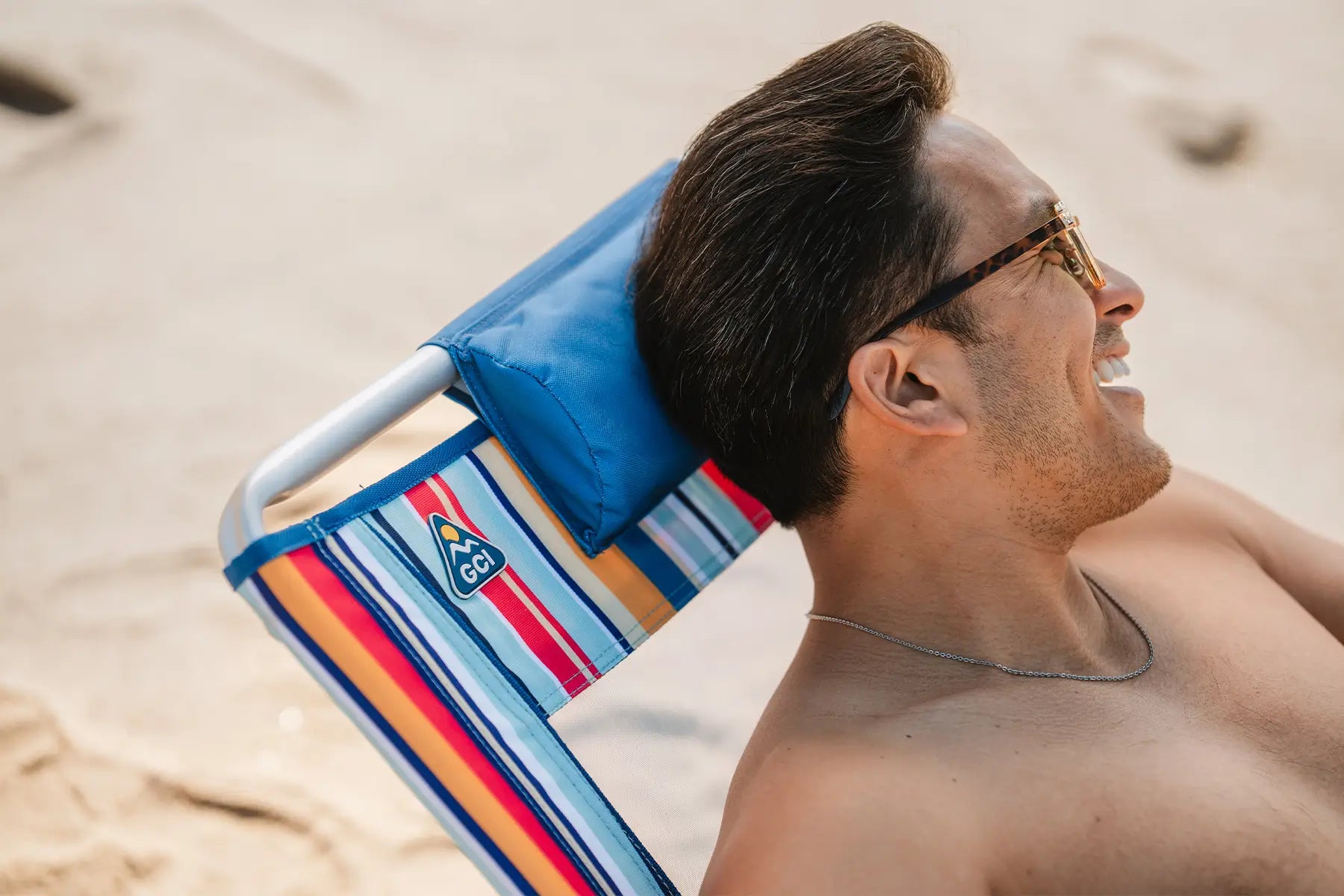 A shirtless man reclined back on a multi stripe Backpack Beach Chair on a beach. 