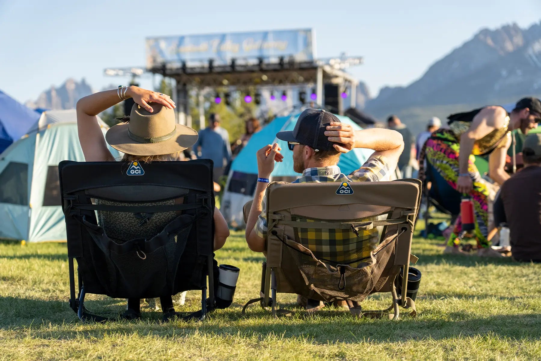 Two people relax in GCI chairs at an outdoor music festival, enjoying the view of the stage with mountains in the background.