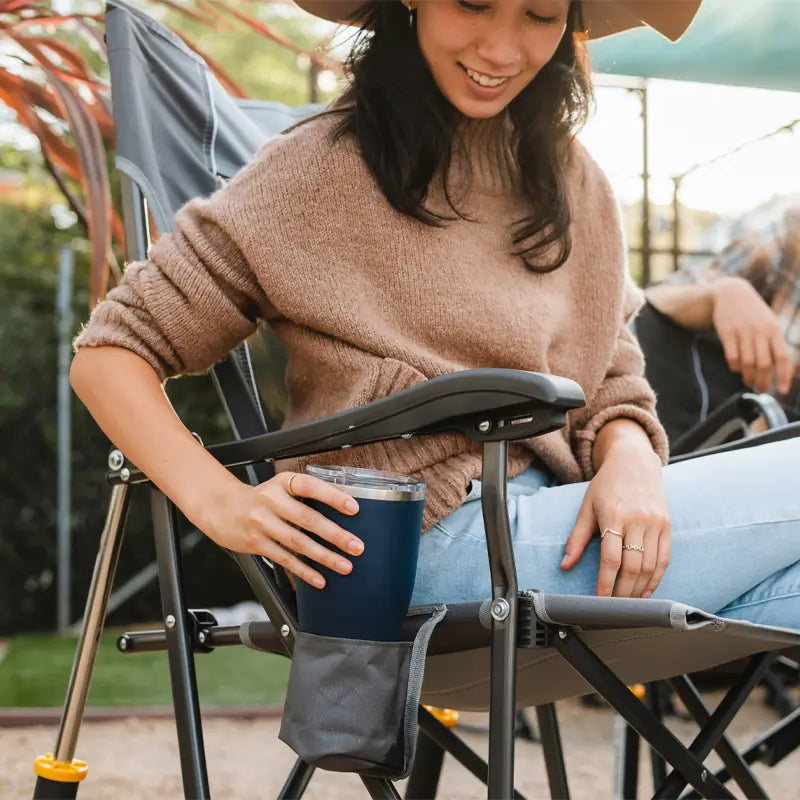 Woman placing a tumbler into the mesh cup holder of a mercury gray RoadTrip Rocker, smiling during an outdoor hangout.
