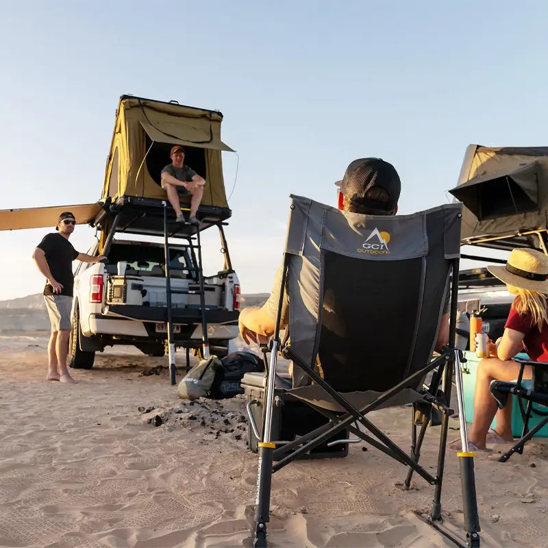Person seated in a mercury gray RoadTrip Rocker on desert sand, facing rooftop tents and friends gathered around a campsite.