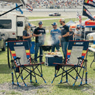 Two NASCAR Roadtrip Rocker chairs in the foreground at an outdoor racetrack event, with people grilling and standing behind them near race cars and tents.