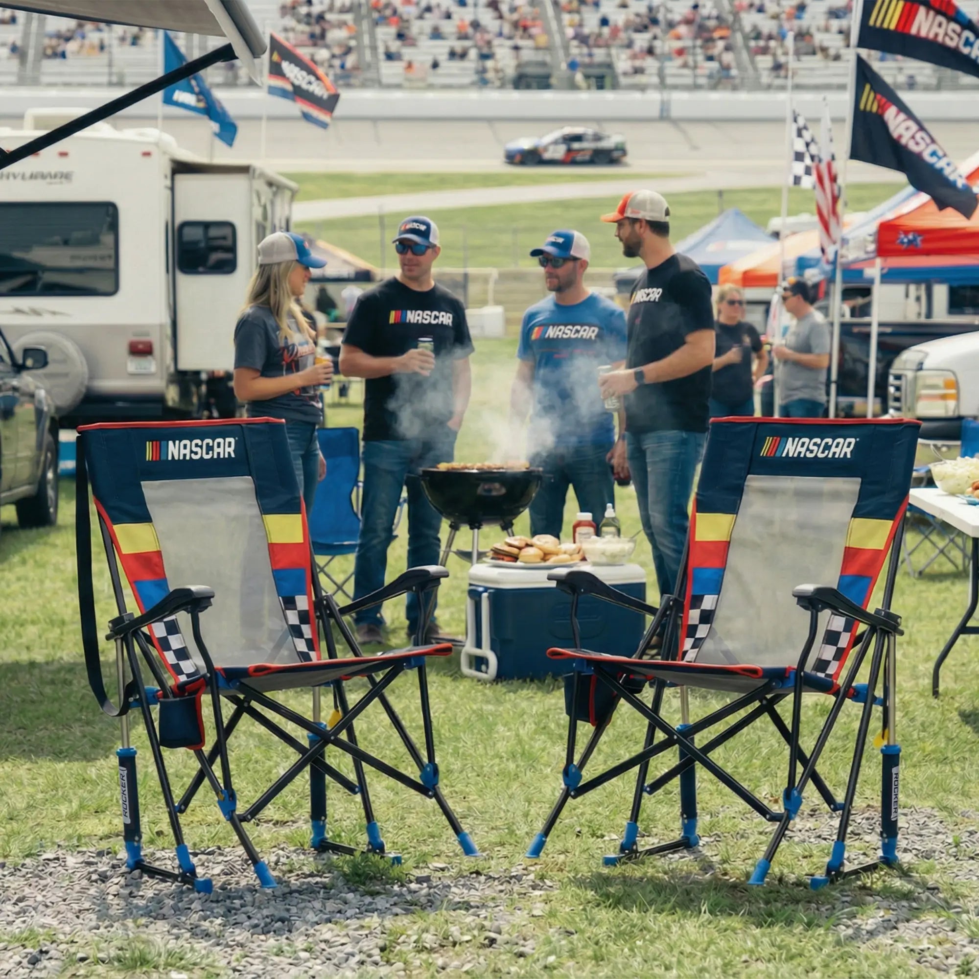 Two NASCAR Roadtrip Rocker chairs in the foreground at an outdoor racetrack event, with people grilling and standing behind them near race cars and tents.