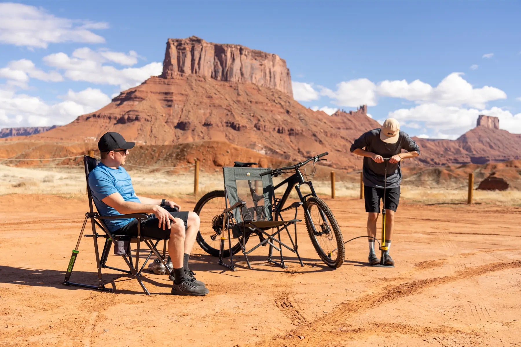 Man sitting in a Roadtrip Rocker in a desert landscape with a bike nearby, while another person pumps air into the tire.