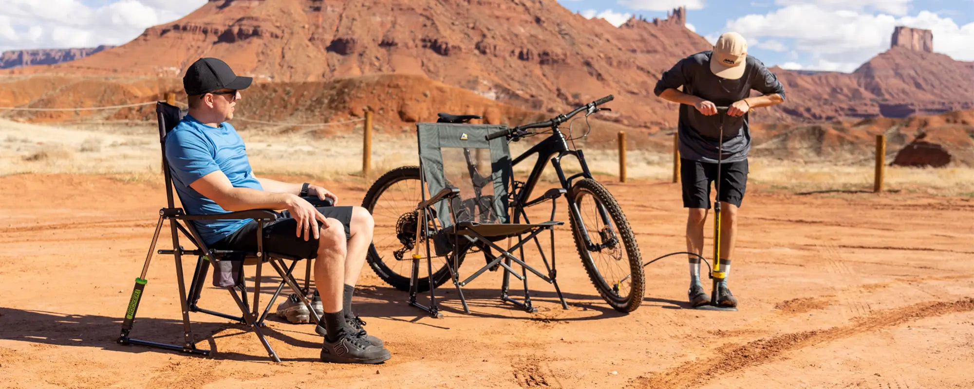 Man sitting in a Roadtrip Rocker in a desert landscape with a bike nearby, while another person pumps air into the tire.