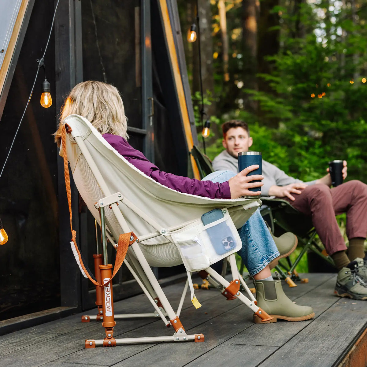A woman sits in a beige and orange Fleece Rocker camp chair on a wooden deck, holding a blue tumbler. A man sits in a matching chair in the background.