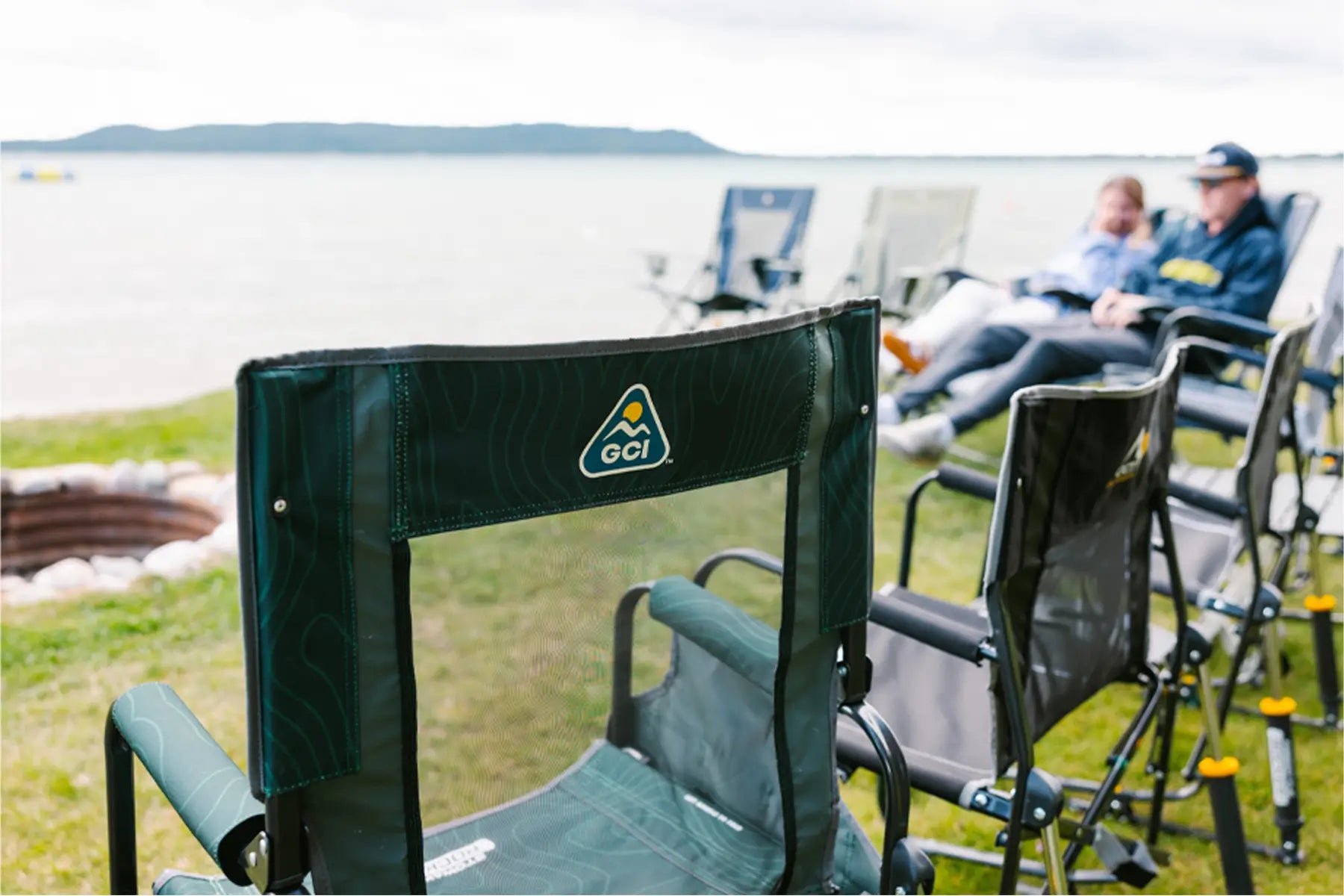 A close-up of a GCI outdoor rocker chair near a fire pit by the lake, with a couple relaxing in the background.