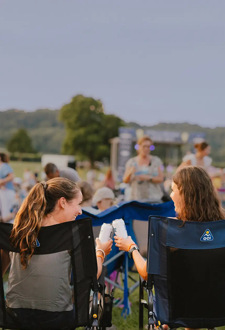 Two women cheering with their drinks at a concert venue while sitting in stowaway rockers. 